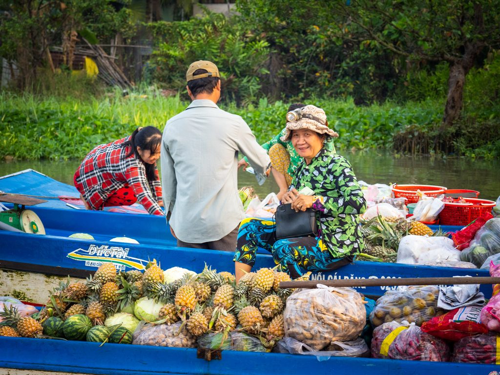 Image of four vendors at Phong Dien floating market on their boats, laden with pineapples and other produce. One of the vendors is looking at the camera and smiling. 