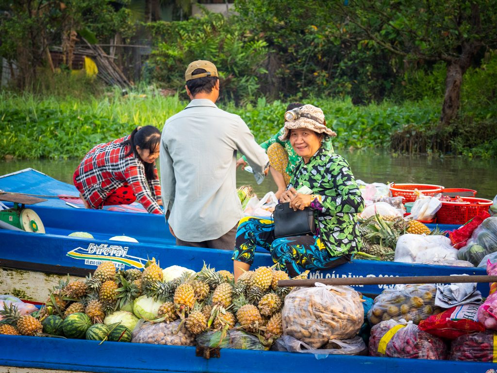 Image of four vendors at Phong Dien floating market on their boats, laden with pineapples and other produce. One of the vendors is looking at the camera and smiling.