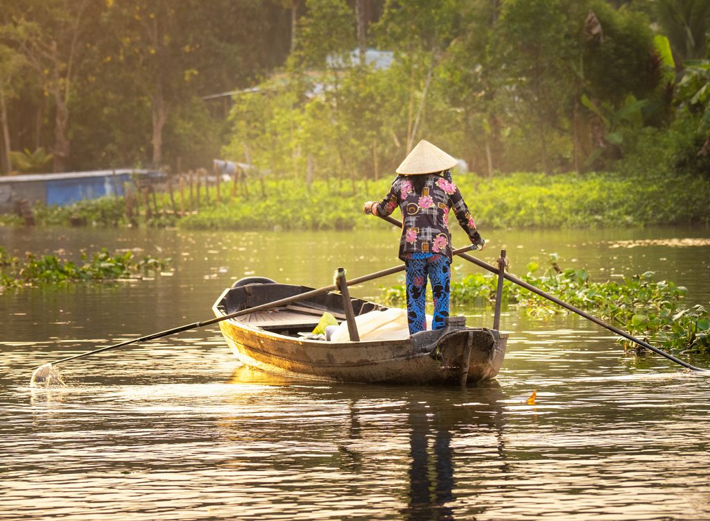 A vendor at Phong Dien floating market rowing her boat and facing away from the camera. The image is bathed in golden light from the early morning sun.